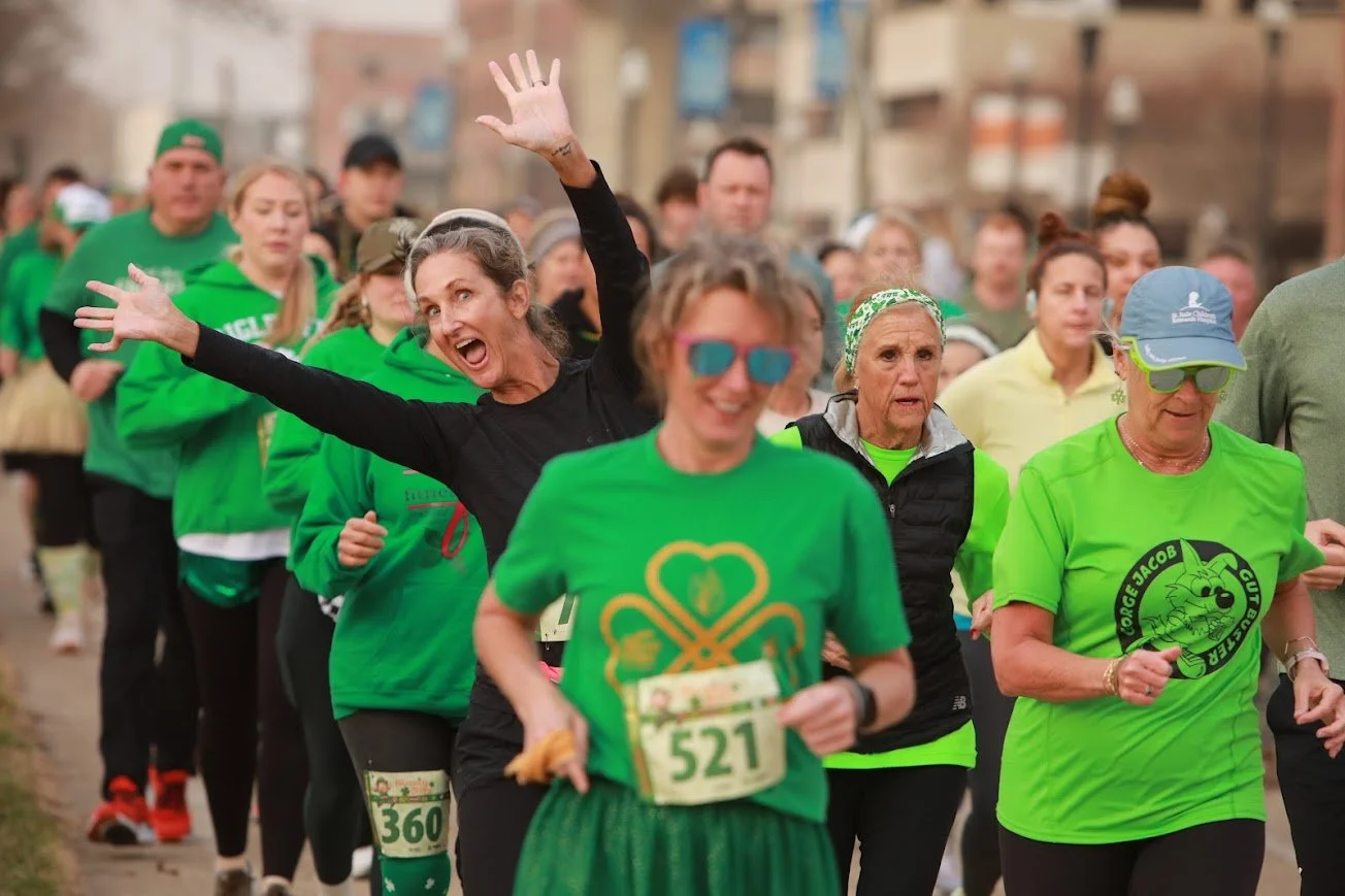 A group of people participating in a race or marathon wearing green clothing, with some smiling and others focusing on running.