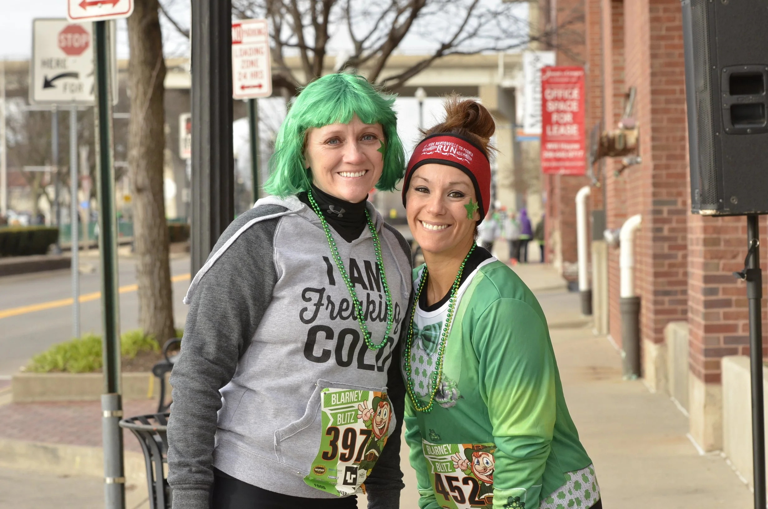 Two women smiling at a street event, both wearing green beads and race bibs, one with green hair and the other in a red headband, celebrating a special occasion or race.