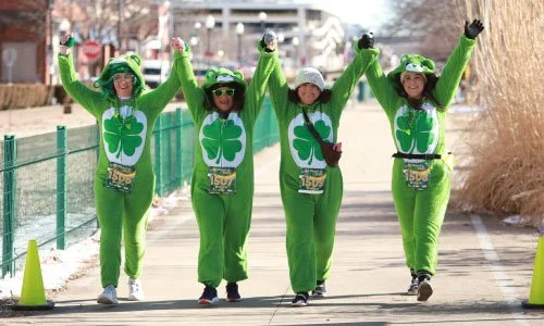 Four people dressed in green shamrock costumes walking outdoors with their hands raised in celebration.