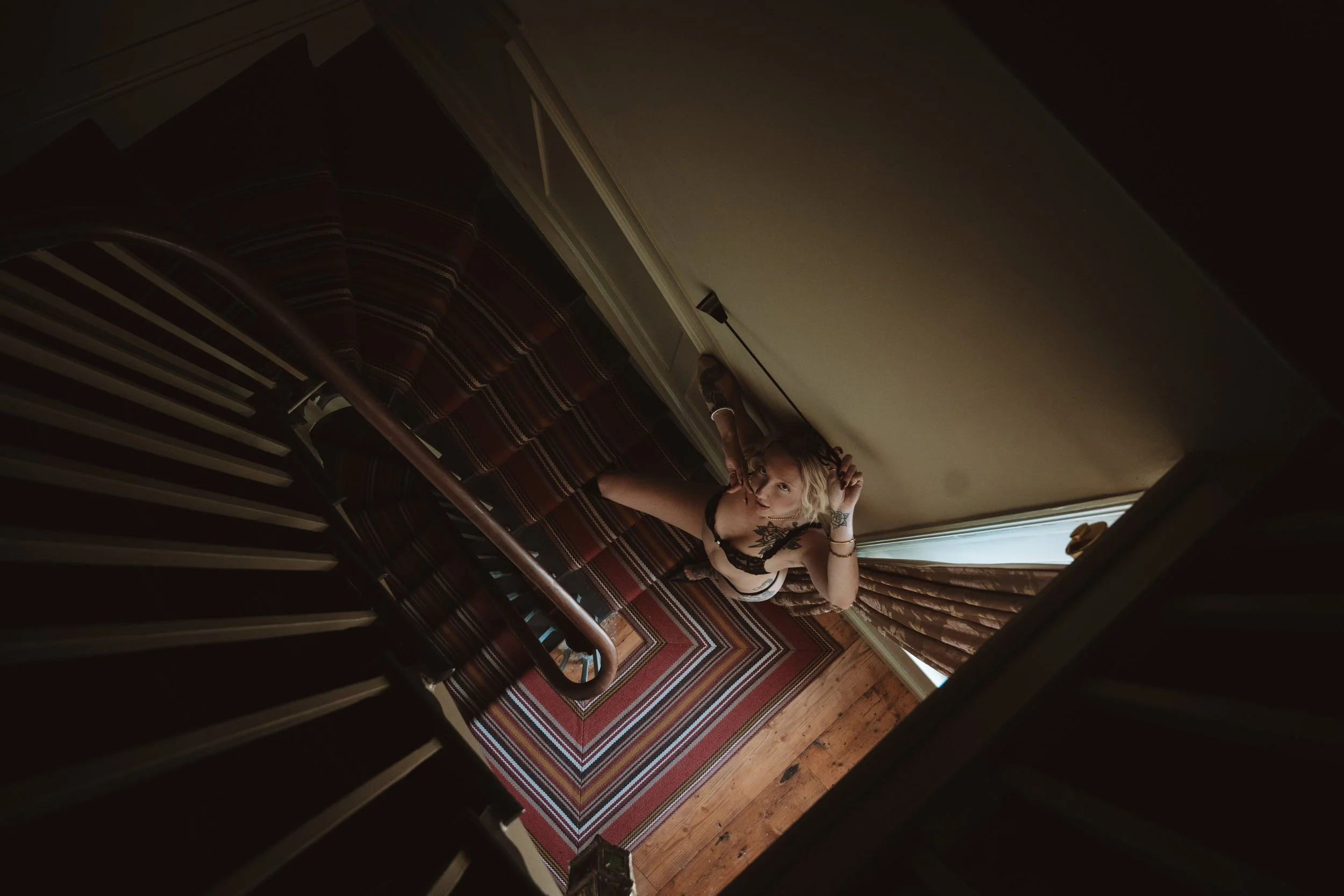 A woman with blonde hair, wearing a black lace top, reclining on a striped sofa in a room with a patterned rug and a wooden floor, with beige curtains and a window in the background.