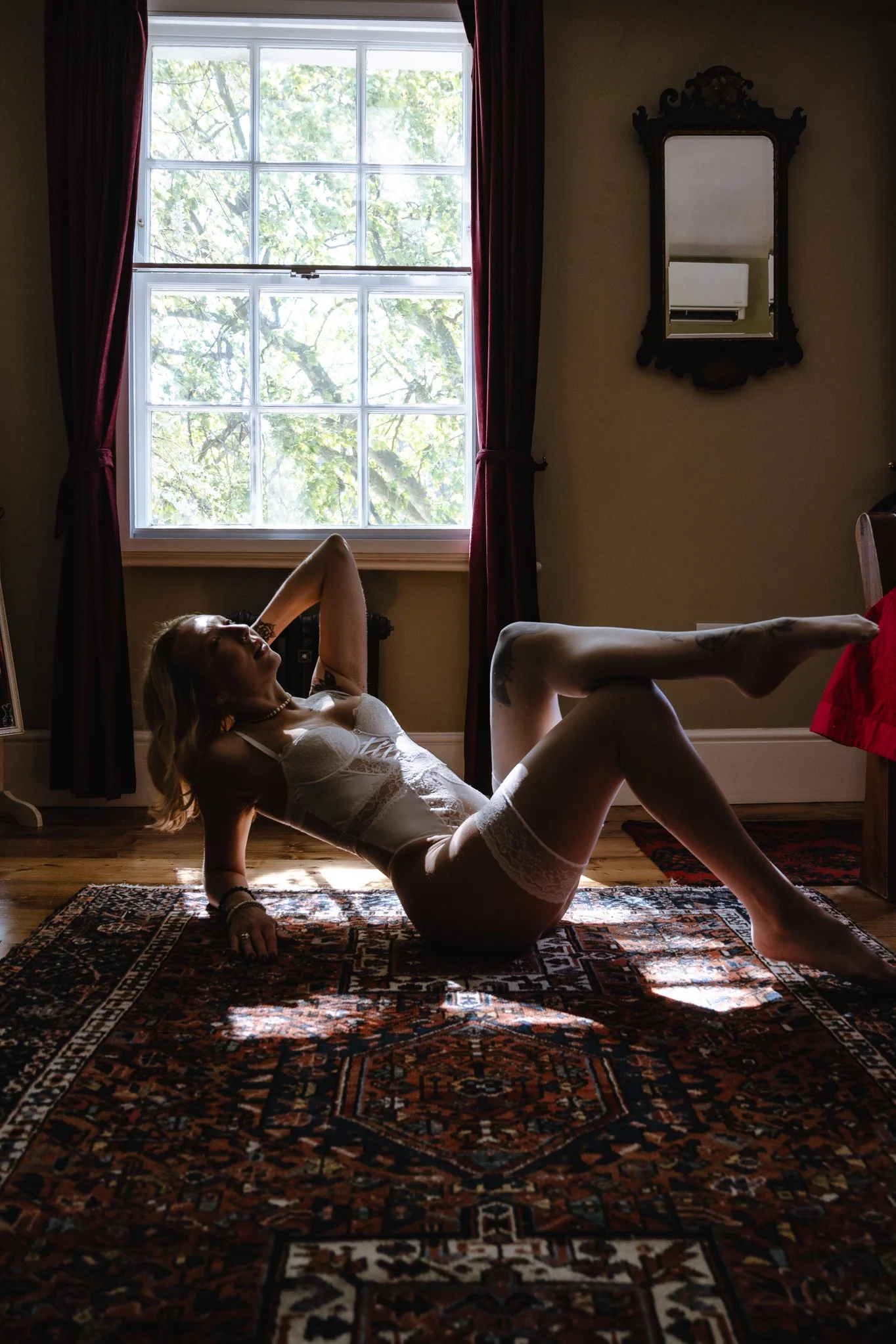 A woman in lingerie lying on a patterned rug in a room with a large window, curtains, and a mirror on the wall.
