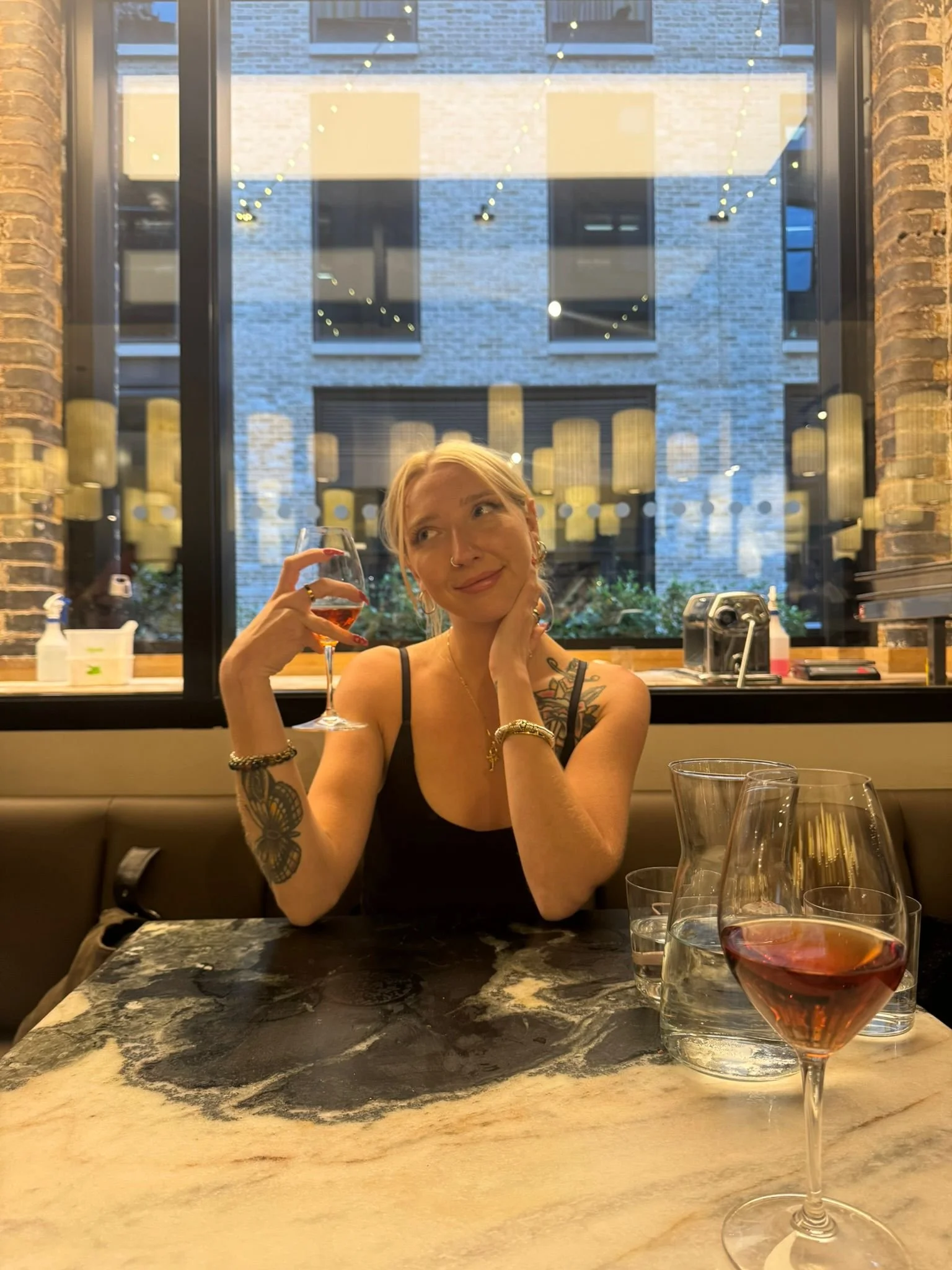 A woman with tattoos on her arm holding a glass of wine and smiling, sitting at a marble table with several glasses of water and wine in front of her, in a restaurant with a large window showing buildings outside.