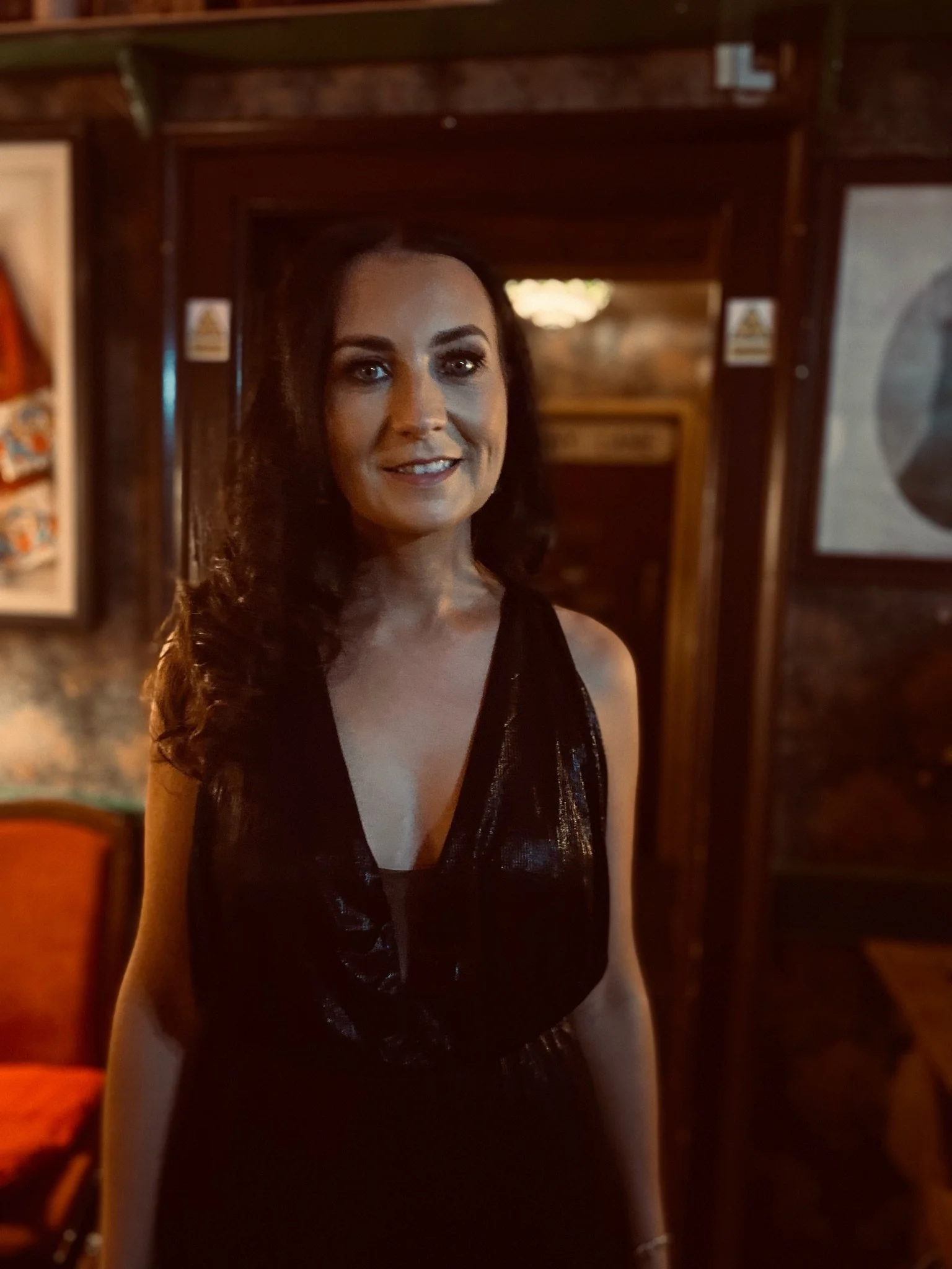 A woman with dark hair, wearing a black shiny sleeveless dress, standing inside a dimly lit wooden restaurant or bar.