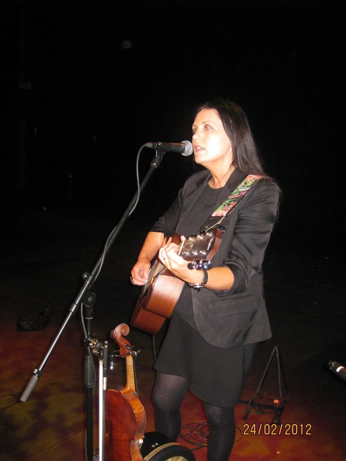 Woman playing guitar and singing into microphone on stage with dark background, wearing black blazer and tights, with a cello on the floor nearby.