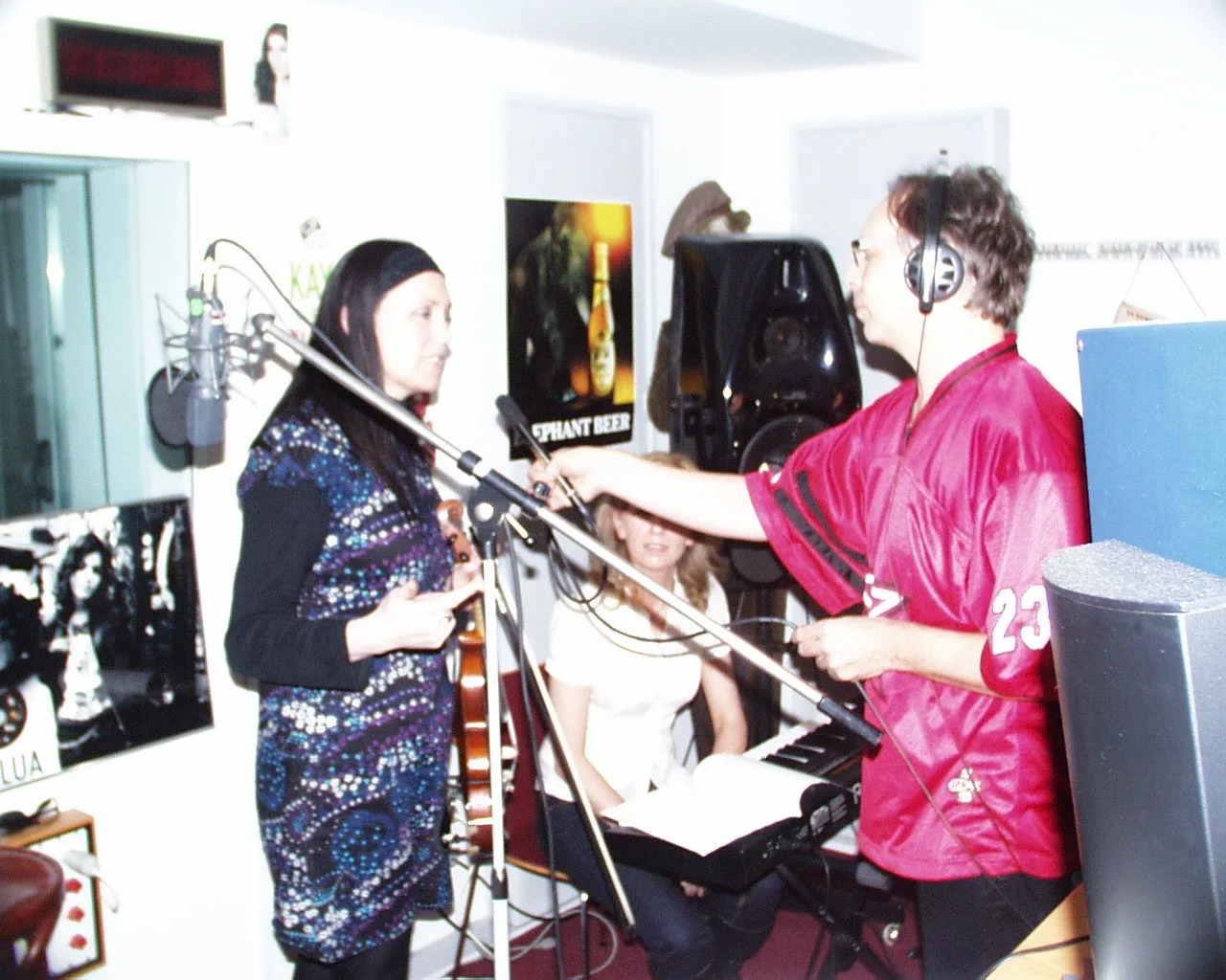 Two musicians performing in a studio, one with a guitar and the other wearing headphones and touching a keyboard, with a woman sitting and watching in the background.
