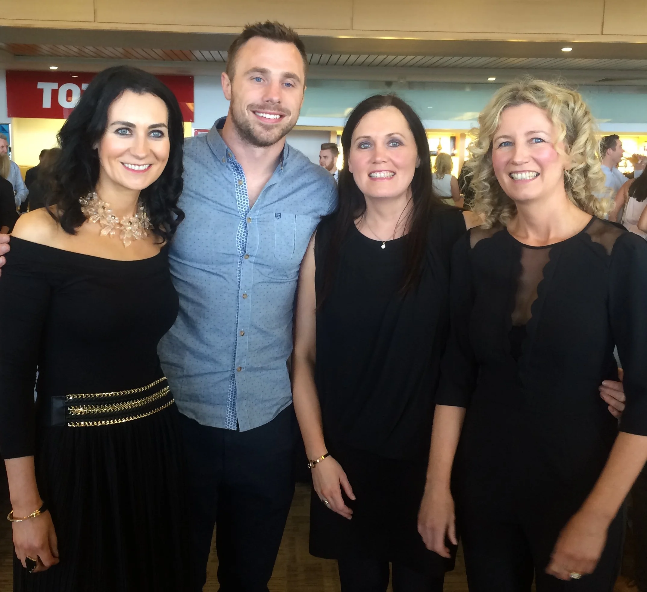 Four people standing together, smiling, in an indoor setting with a wood ceiling and a red "TO" sign in the background.