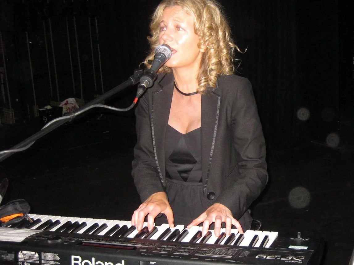 A woman with curly blonde hair singing and playing a keyboard on stage.