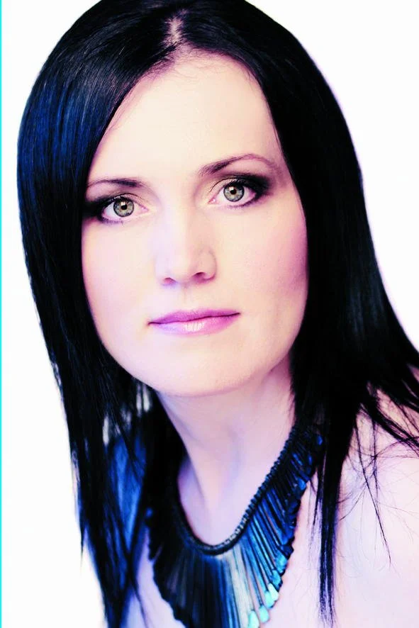 A woman with straight black hair and gray eyes, wearing a black beaded necklace, looking directly at the camera with a neutral expression.