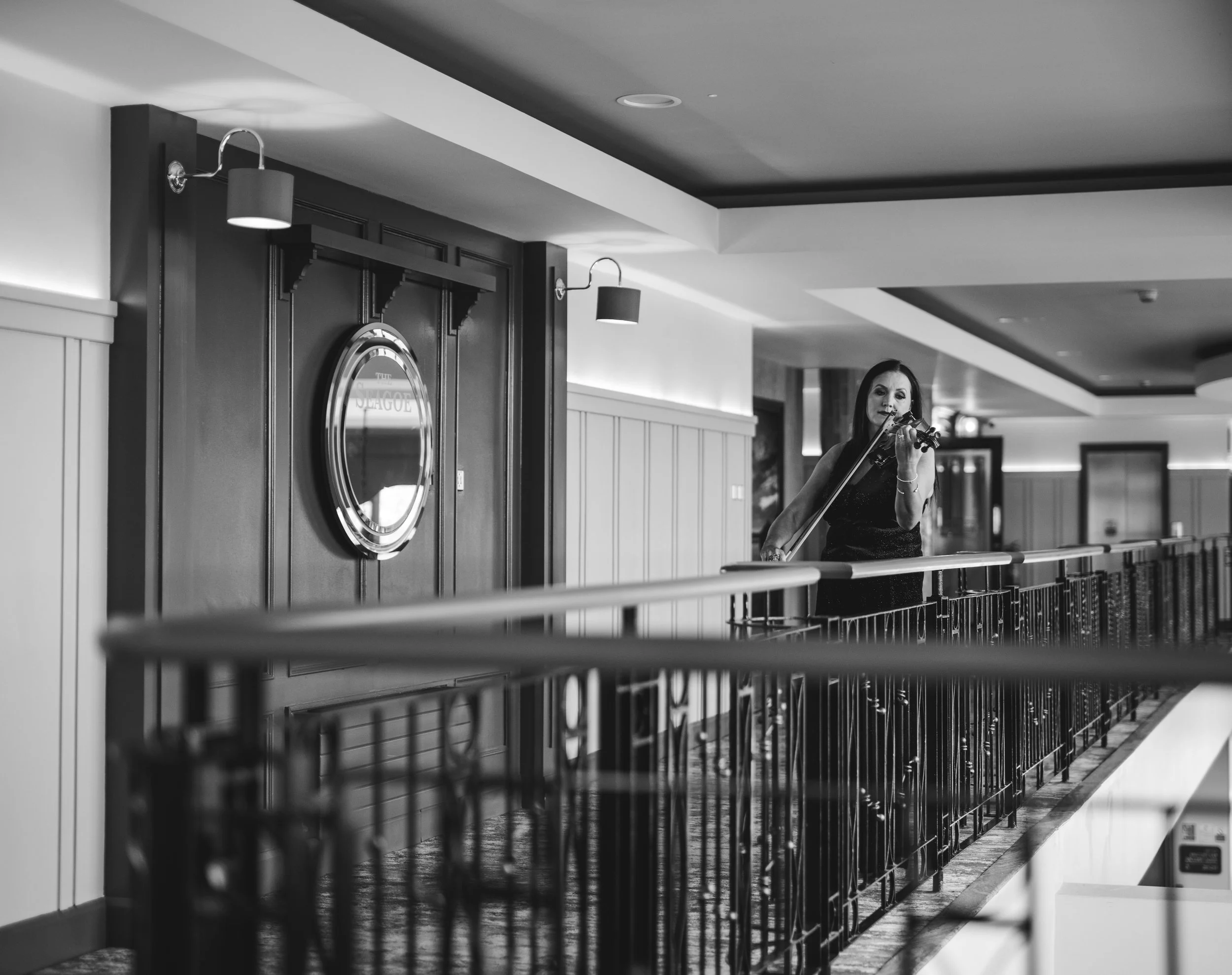 A woman playing the violin on a balcony inside a building with wood-paneled walls and modern light fixtures.