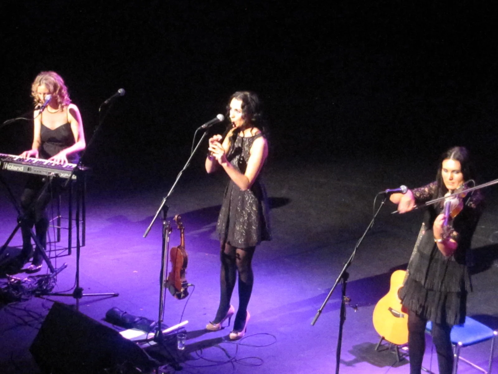 Three female musicians performing on stage. The woman in the center is singing into a microphone, while the woman on the right is playing a violin, and the woman on the left is playing a keyboard.