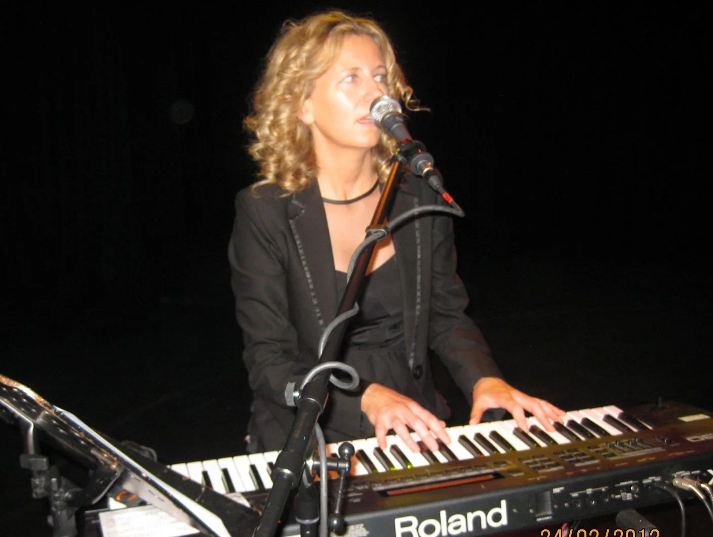 A woman with curly blonde hair wearing a black blazer, playing a keyboard on stage with a microphone in front of her.