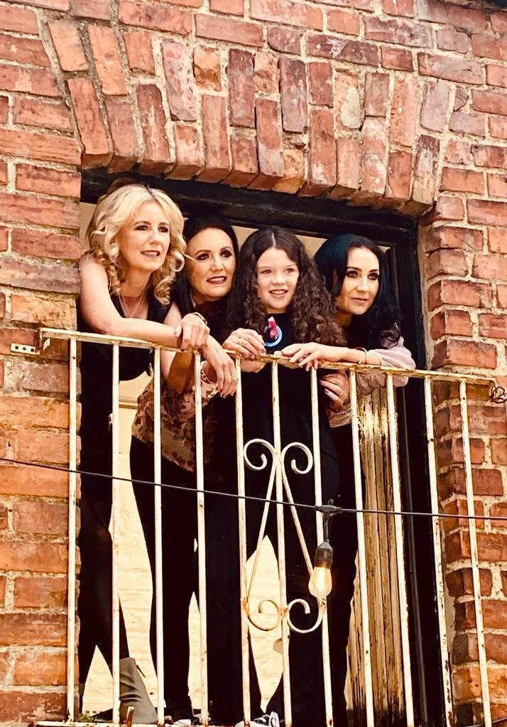 Four women and a young girl leaning on a balcony railing of a brick building, smiling and looking out.