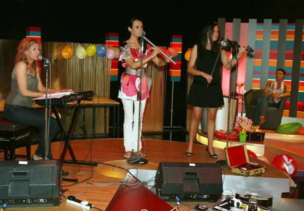 Three women performing a music act on stage with a keyboard, flute, and violin, in a colorful, decorated indoor setting, with a woman sitting on a couch in the background.