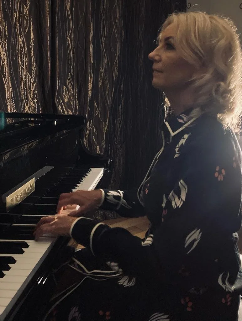 A woman with blonde curly hair playing a black grand piano indoors, wearing a black blouse with white and pink floral patterns.