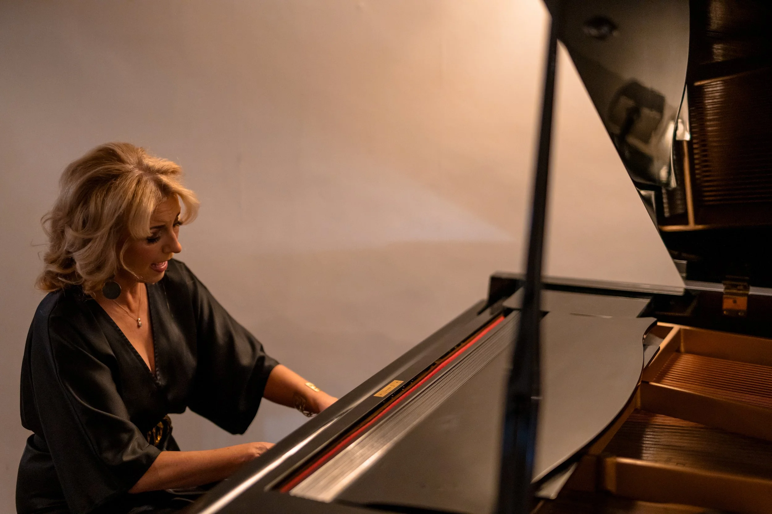 A woman with blonde hair playing a grand piano, wearing a black satin top, in what appears to be a performance or practice setting.