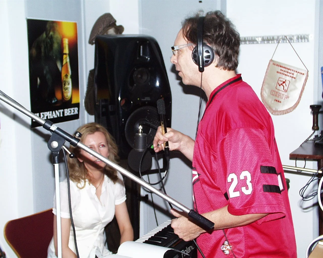 A man wearing a red sports jersey and headphones is singing or speaking into a microphone, standing next to a woman sitting at a keyboard in a room with a poster of Elephant Beer and a speaker in the background.