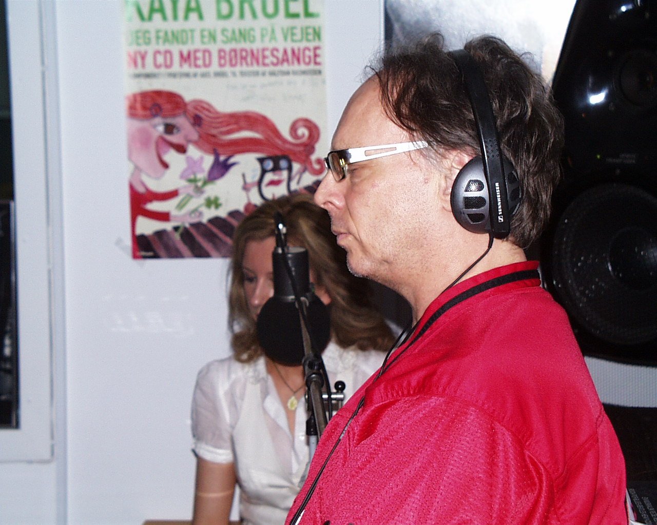 A man wearing glasses and headphones, with eyes closed, standing in front of a microphone, and a woman with a microphone in front of her, in a recording studio with a poster in the background.