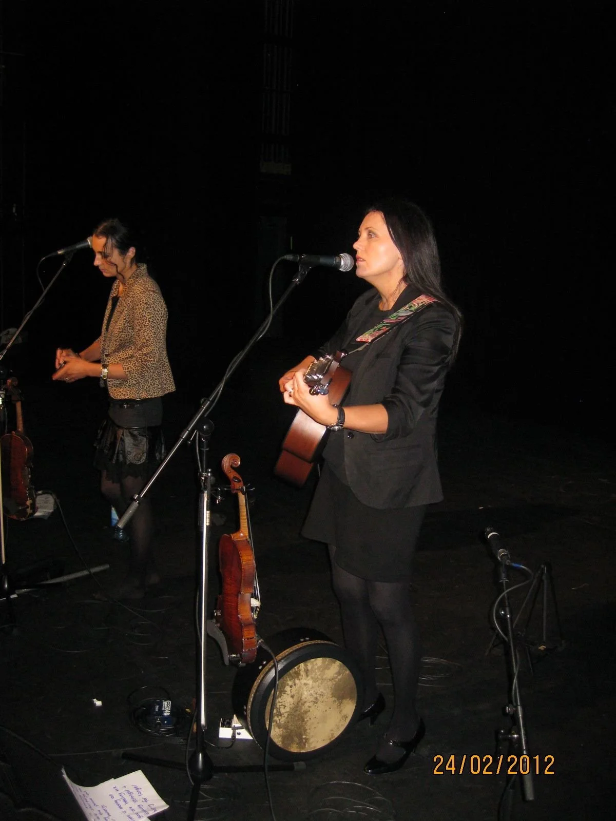 Two women performing on stage with microphones, one playing guitar and the other with a violin, against a black background, with the date 24/02/2012 displayed.