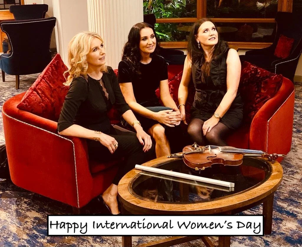 Three women sitting on a red velvet couch with a violin and a bow on the table in front of them, celebrating International Women's Day.