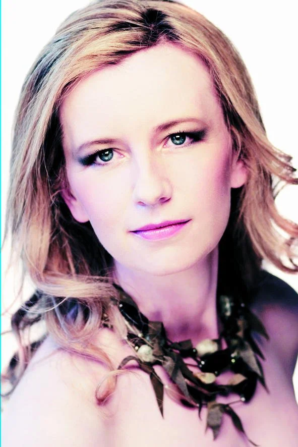 Portrait of a woman with long wavy blonde hair, fair skin, and blue eyes, wearing a dark necklace with leaf designs, against a white background.