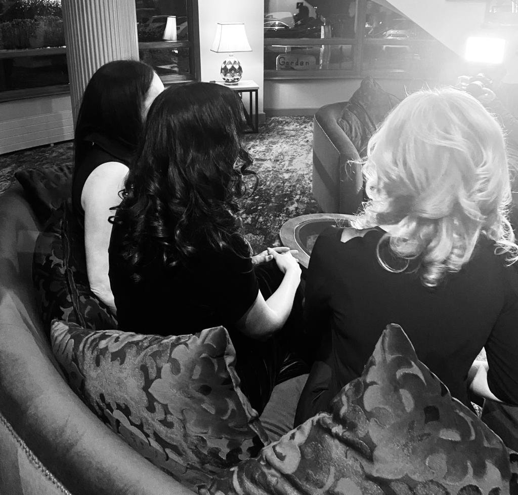 Three women sitting on a couch in a living room, facing away from the camera. Two have dark hair, one has light, and all are dressed in dark clothing. They are engaged in conversation, with a table and a lamp in the background.