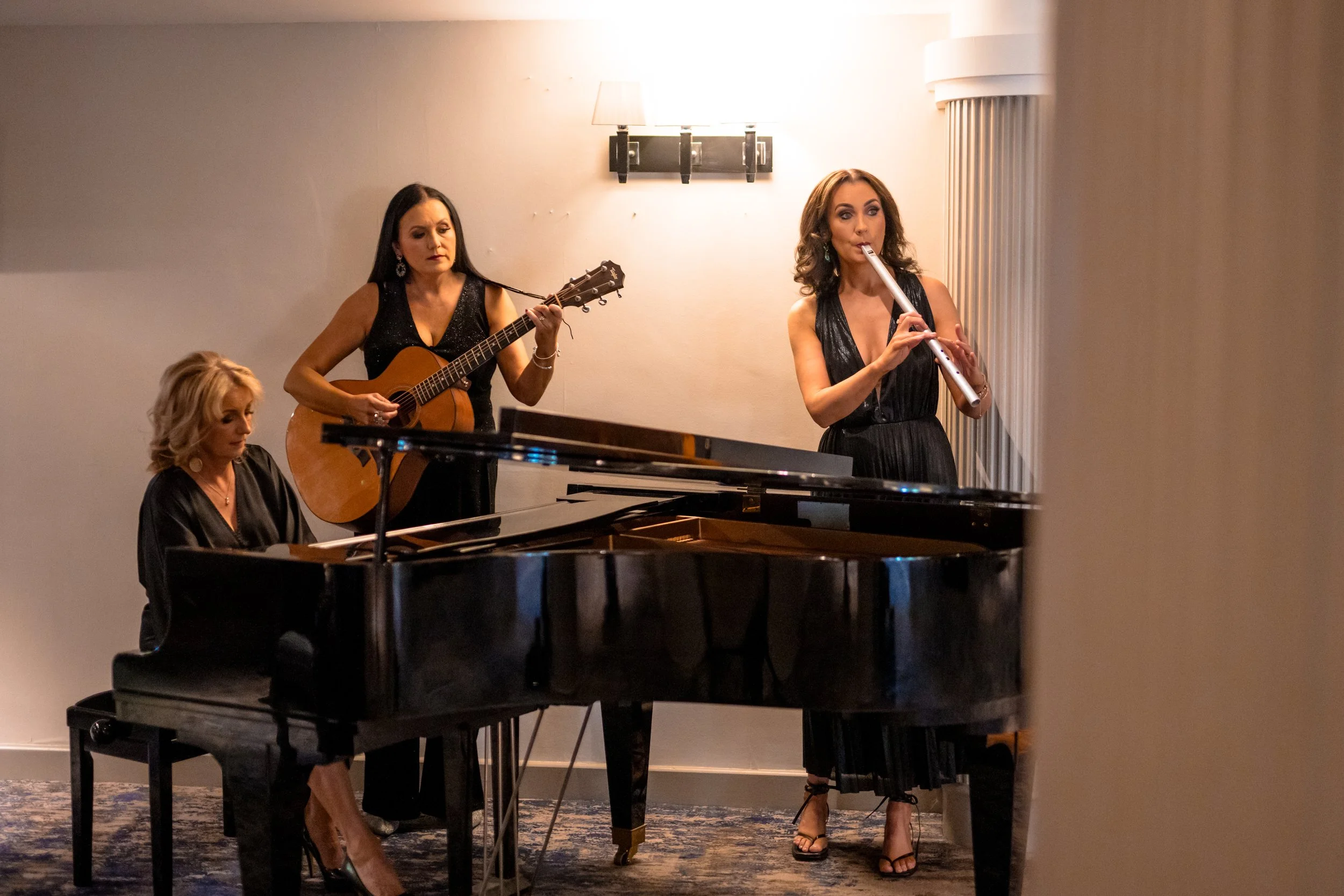 Four women performing music together: one playing the piano, one playing the guitar, one singing into a microphone, and one playing the flute in a cozy indoor setting.