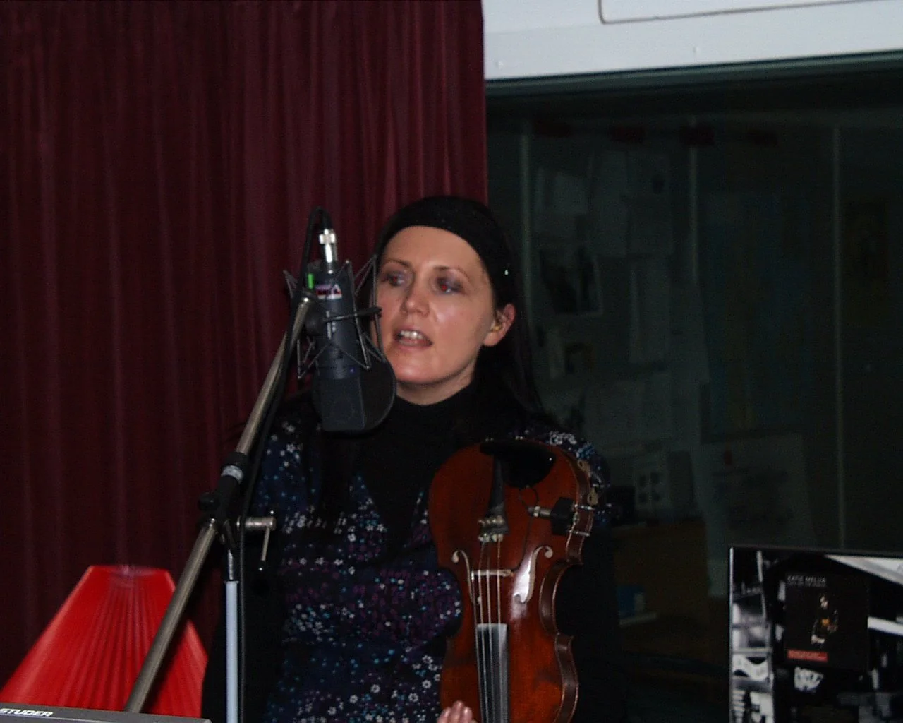 A woman with dark hair, wearing a black clothing with sparkles, singing into a microphone while holding a violin, in a recording studio with red curtains and soundproof walls.