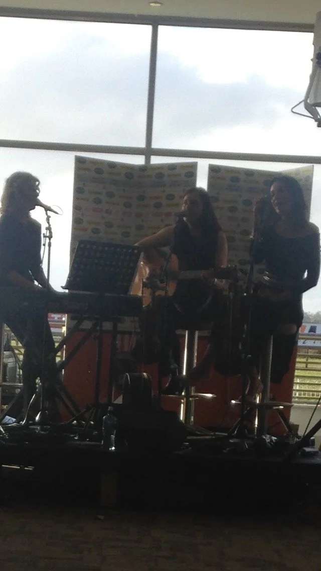 Three women performing on a stage with musical instruments in front of large windows, in an indoor venue during the daytime.