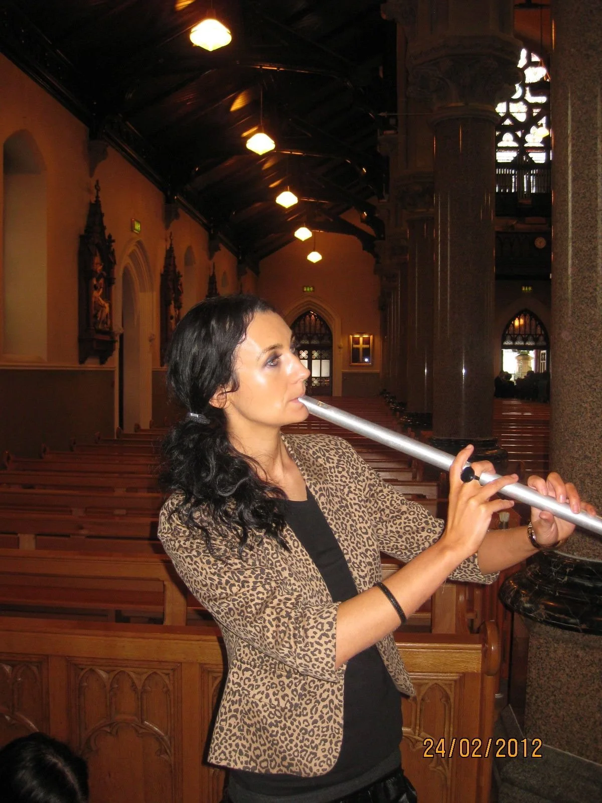 A woman with dark, curly hair and a leopard-print blazer is playing a silver flute inside a church or cathedral with wooden pews and high ceilings.