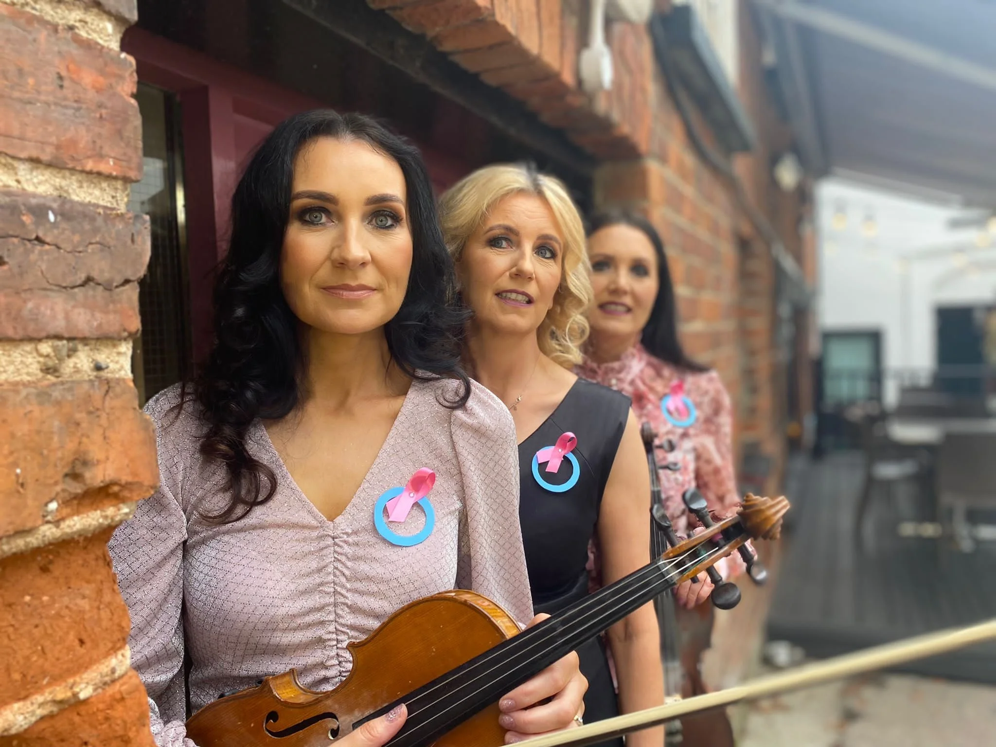 Three women standing outdoors in front of a brick building, each with a pink and blue awareness ribbon badge. The woman in front has dark hair and is holding a violin, the second woman has blonde hair, and the third has dark hair and is holding a bow