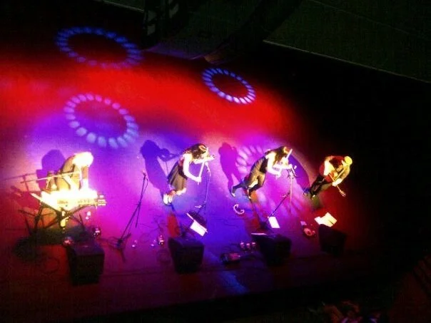 Four people perform on a stage with colorful lighting and circular patterns on the floor; sounds equipment is also visible.