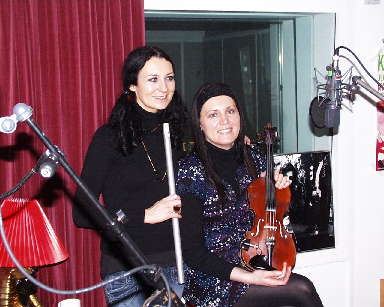 Two women in a recording studio, one holding a flute and the other holding a violin, with microphones and sound equipment around them.