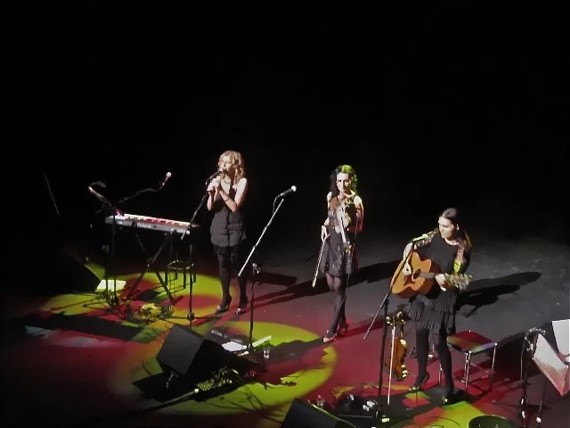 Three female musicians performing on stage with a keyboard, a microphone, and a guitar, under stage lighting.