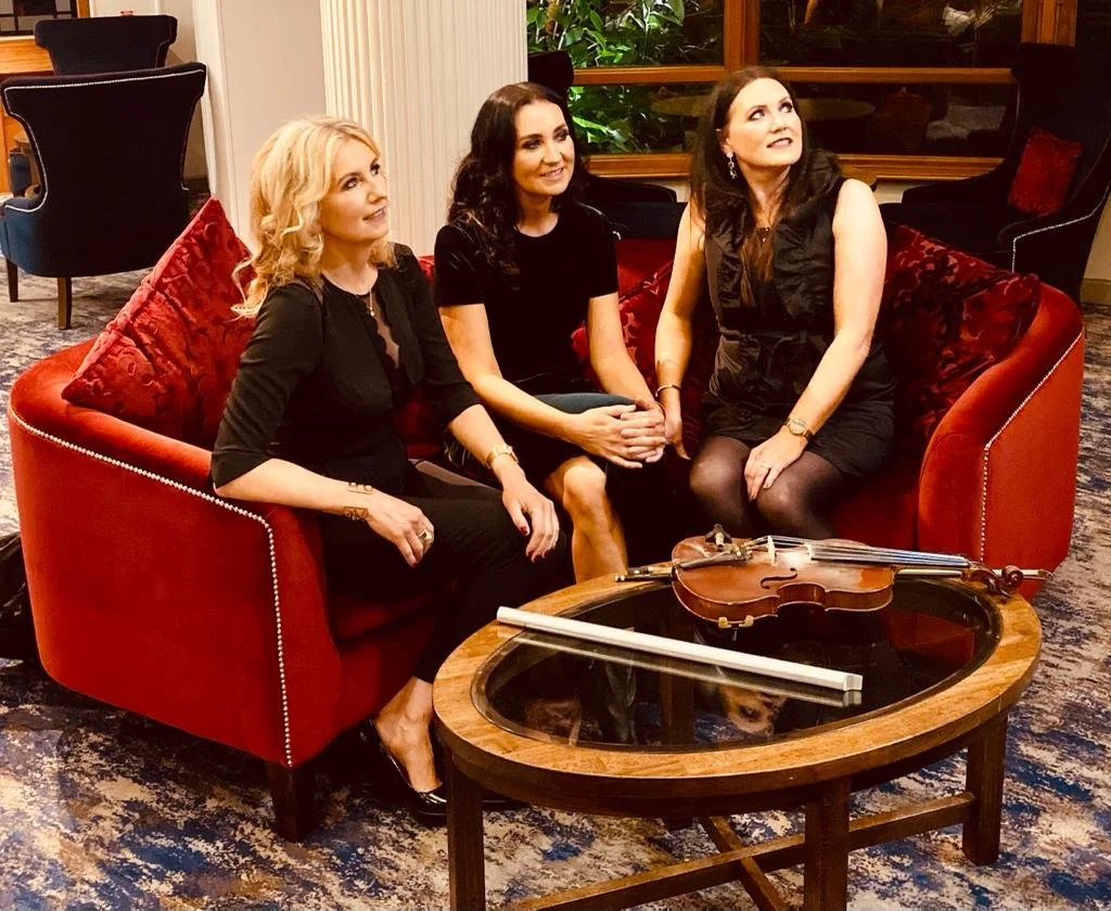 Three women sitting on a red velvet sofa in a room with side chairs and wooden window frames, with musical instruments - a violin and a bow on a round coffee table in front of them.