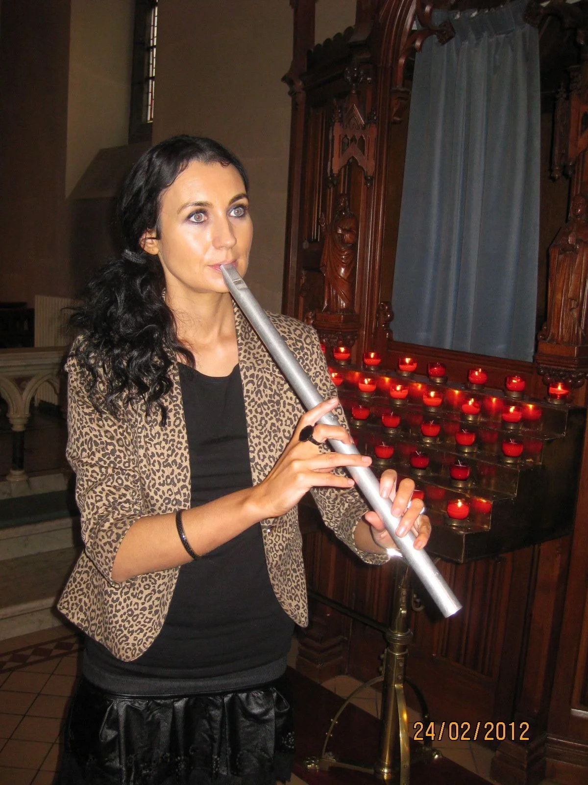 A woman with dark, curly hair playing a silver flute in a church, with lit red candles behind her.