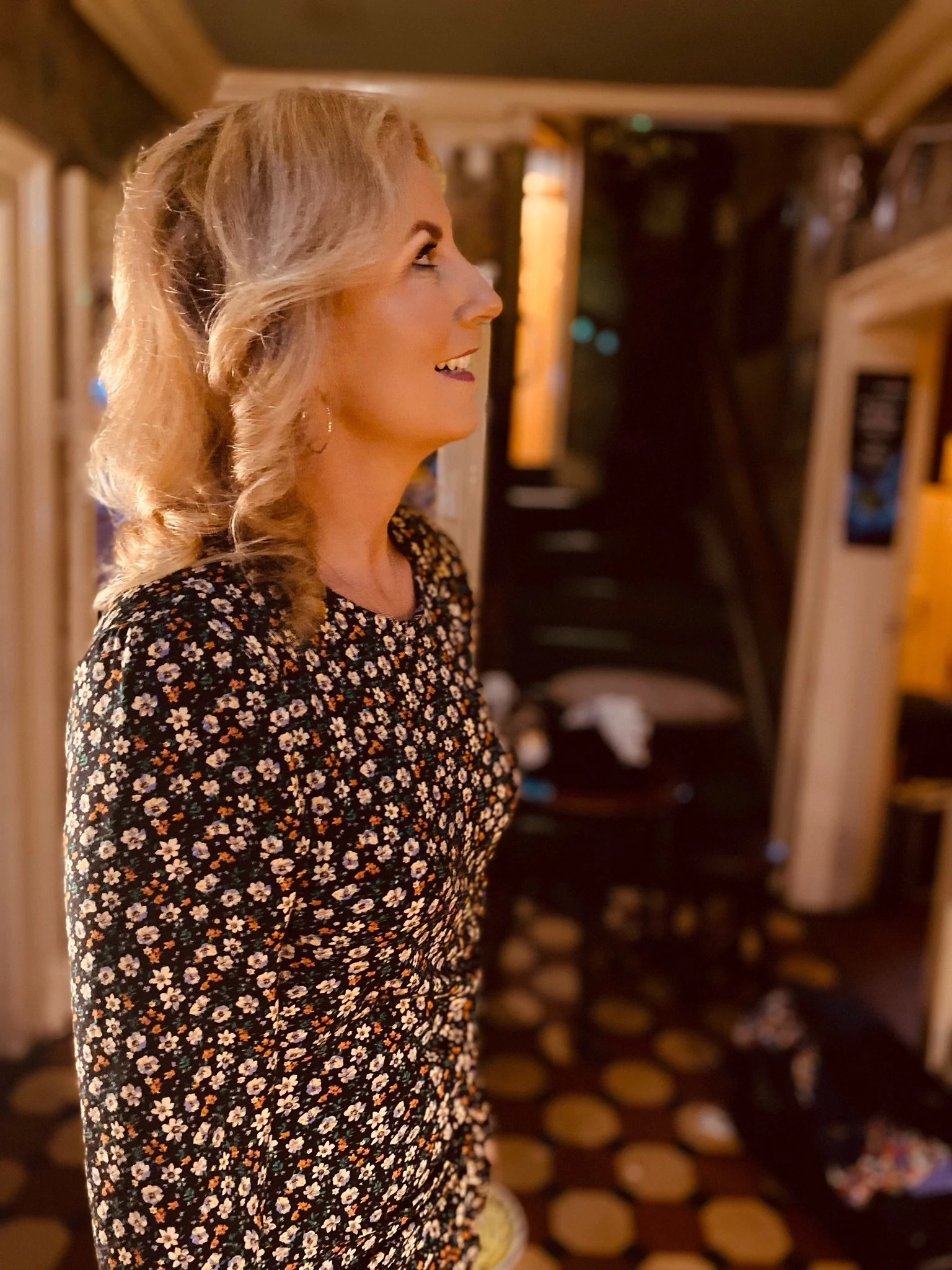 A woman with blonde wavy hair styled to one side, smiling and looking to her right. She is wearing a black floral dress with small white and orange flowers. The background shows a cozy indoor setting with warm lighting, a black chair, and wood-panele