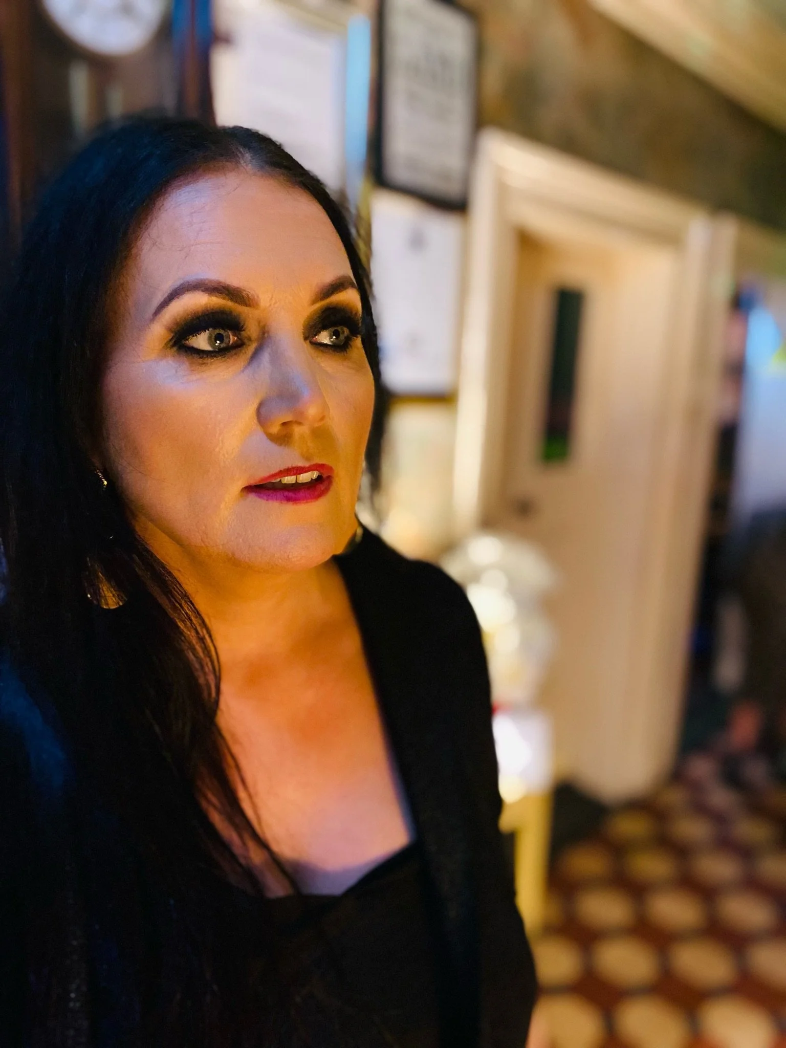 A woman with black hair and makeup looking to the left, standing indoors with certificates or framed items on the wall behind her.
