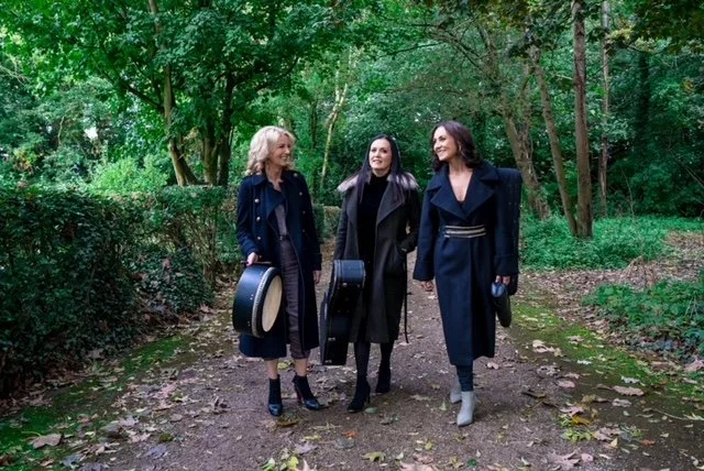 Three women walking together in a wooded park, engaged in conversation, dressed in stylish coats, carrying musical instrument cases.