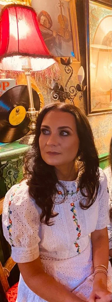 A woman with dark curly hair and wearing a white embroidered dress sits in a decorated room with vintage art and a red lampshade.