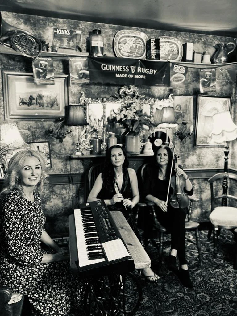 Three women in a cozy, vintage-style room with a piano, a violin, and a flute, surrounded by framed pictures and decorative items, posing for a photo.