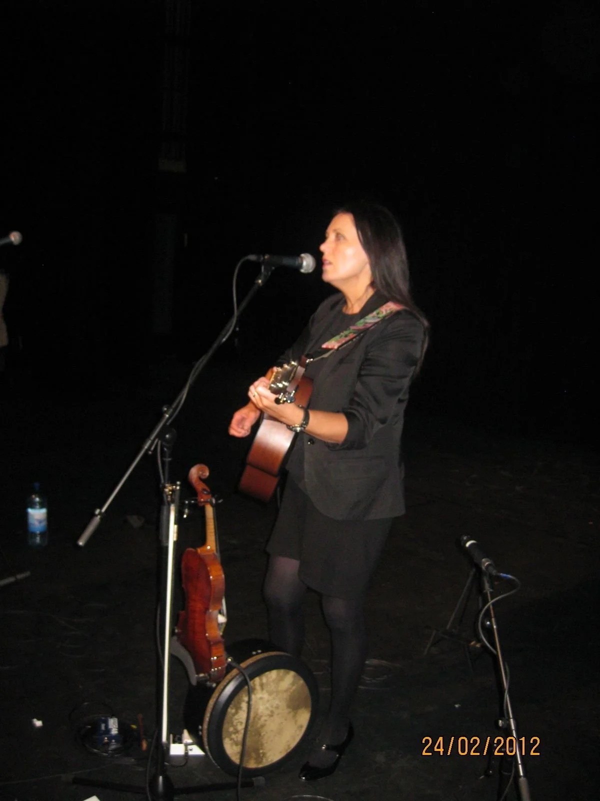 A woman dressed in black with long dark hair, playing a guitar and singing into a microphone on a stage, with a second guitar and a tambourine on the floor, and a bottle of water nearby, in a dark performance setting, dated February 24, 2012.