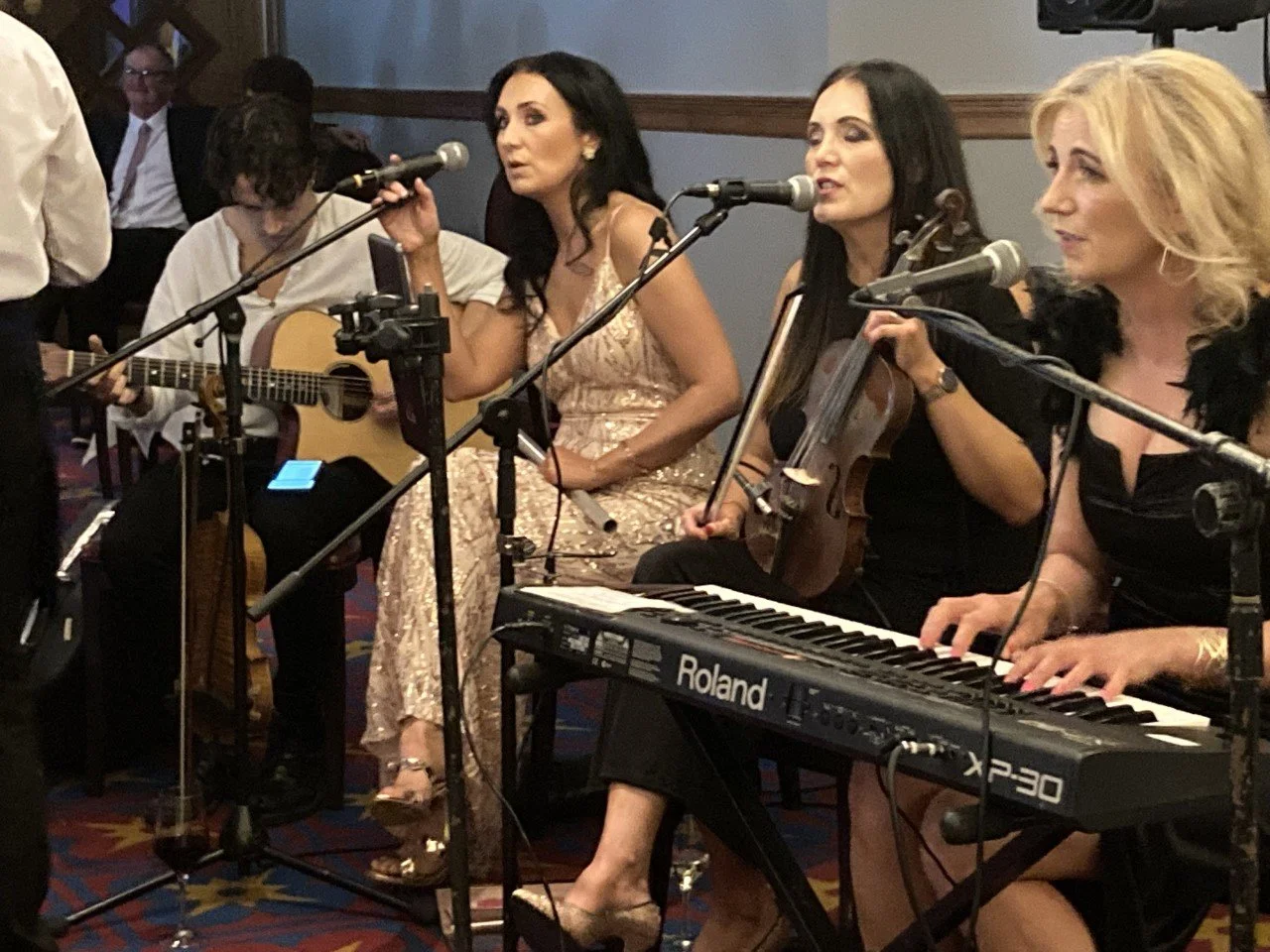 Four women performing music with a guitar, keyboard, and violin at an indoor event.