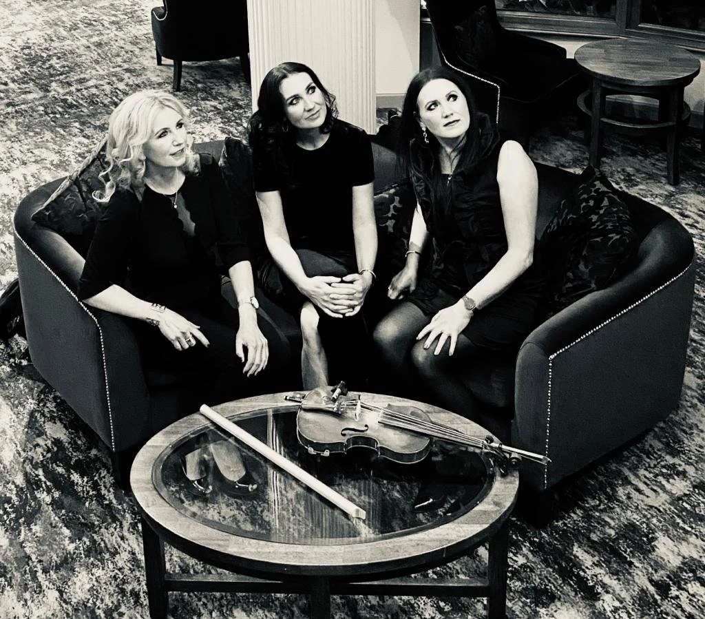 Three women sitting on a dark couch, with a violin and bow on a glass-topped coffee table in front of them, in a living room with patterned carpet.