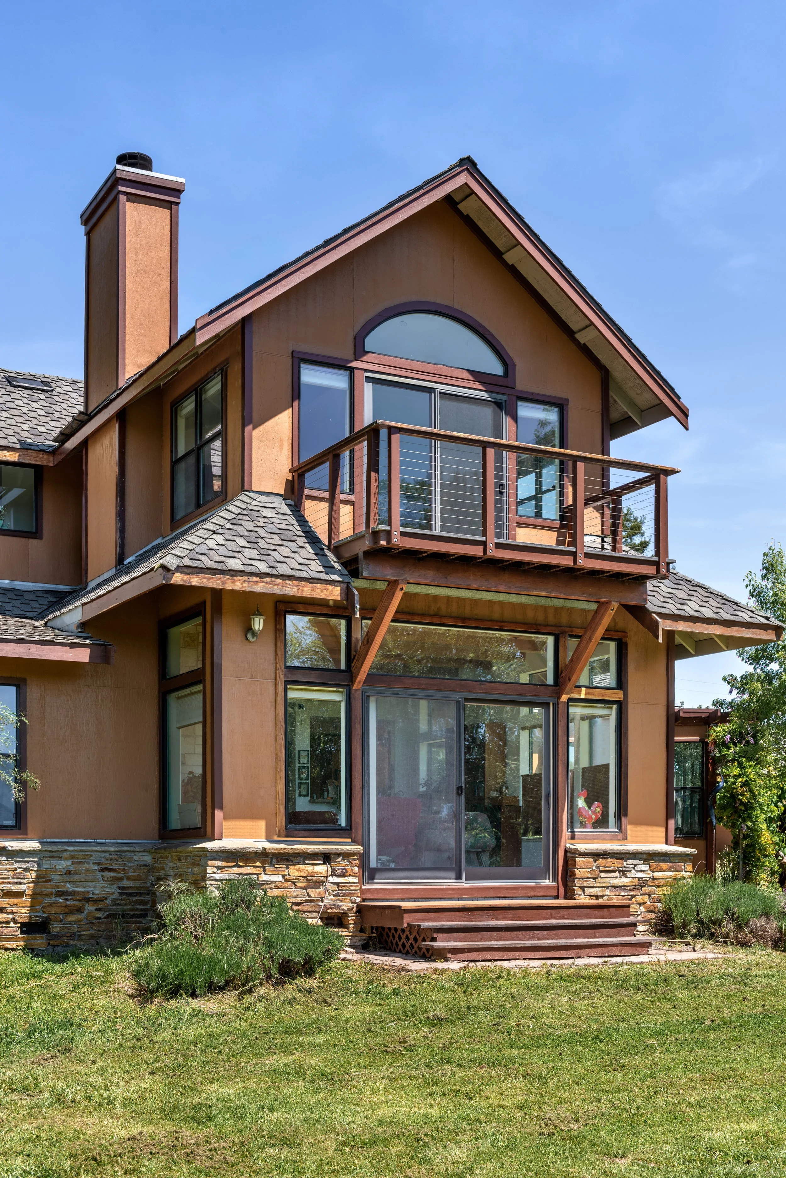 A multi-story house with large glass sliding doors leading to a wooden balcony, brick chimney, and a well-maintained yard with grass and small bushes.