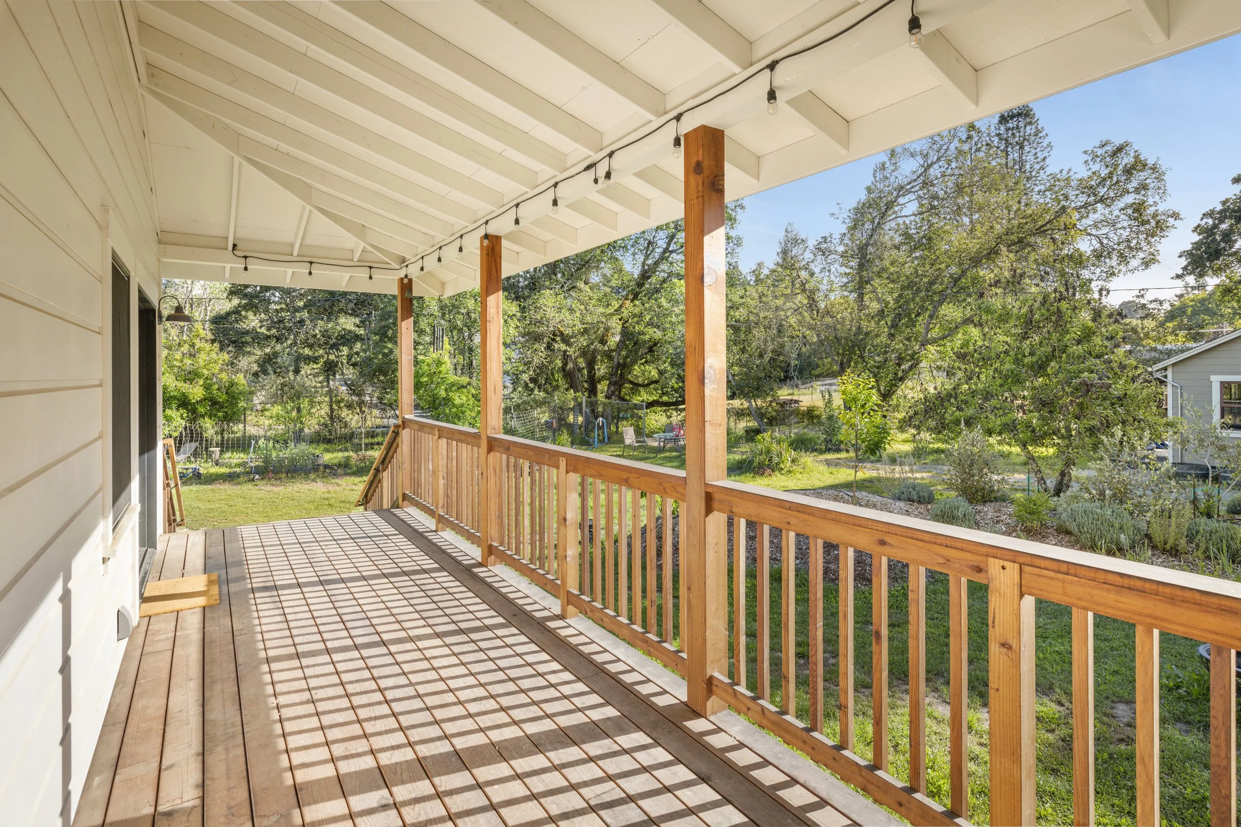 Wooden porch with railing, string lights hanging under a white roof, surrounding greenery and trees in the background.