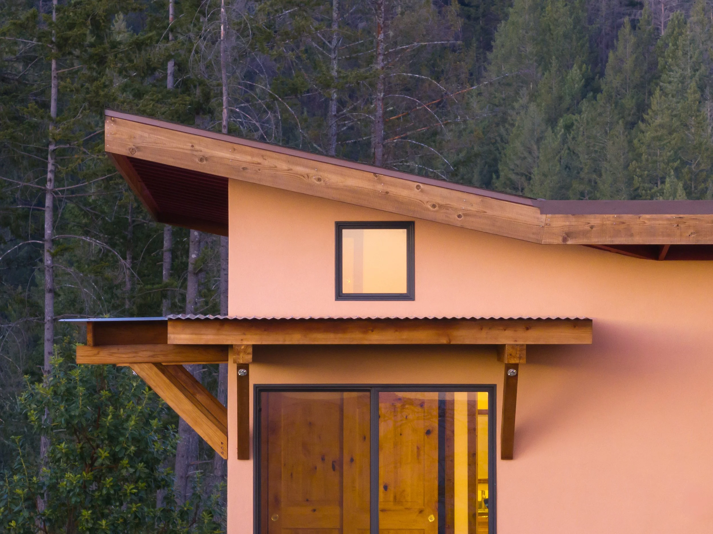 A modern house with yellow exterior walls, large windows, and a sloped roof, set against a backdrop of a forested hillside.