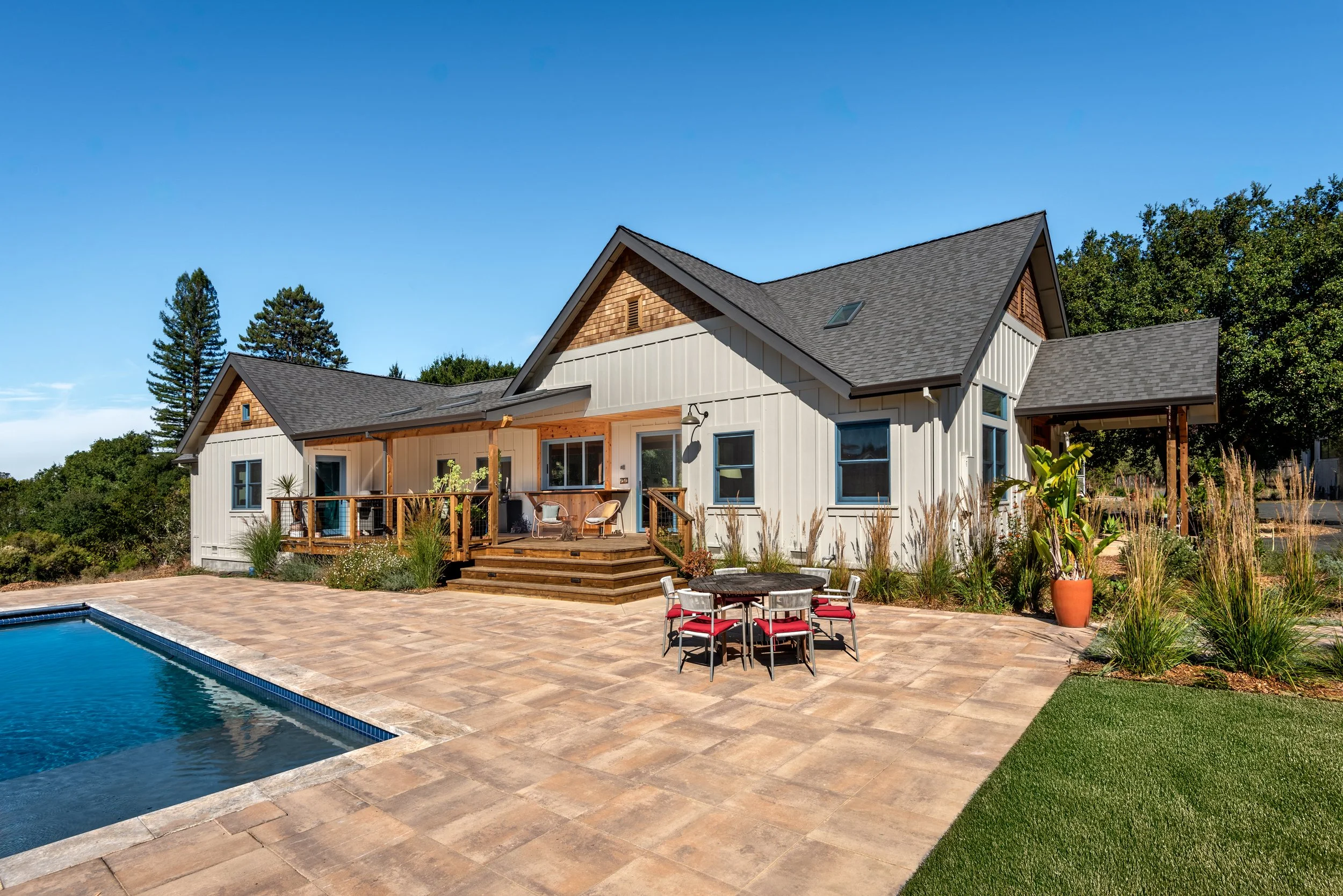 A modern house with a swimming pool in the backyard, surrounded by a stone patio, outdoor furniture, and landscaped plants under a clear blue sky.