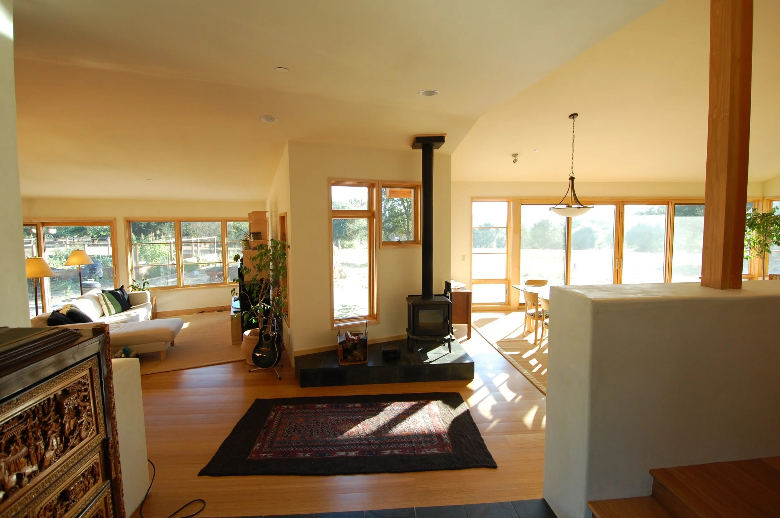 Living room with large windows, a wood stove, a sofa, and potted plants, illuminated by natural sunlight.