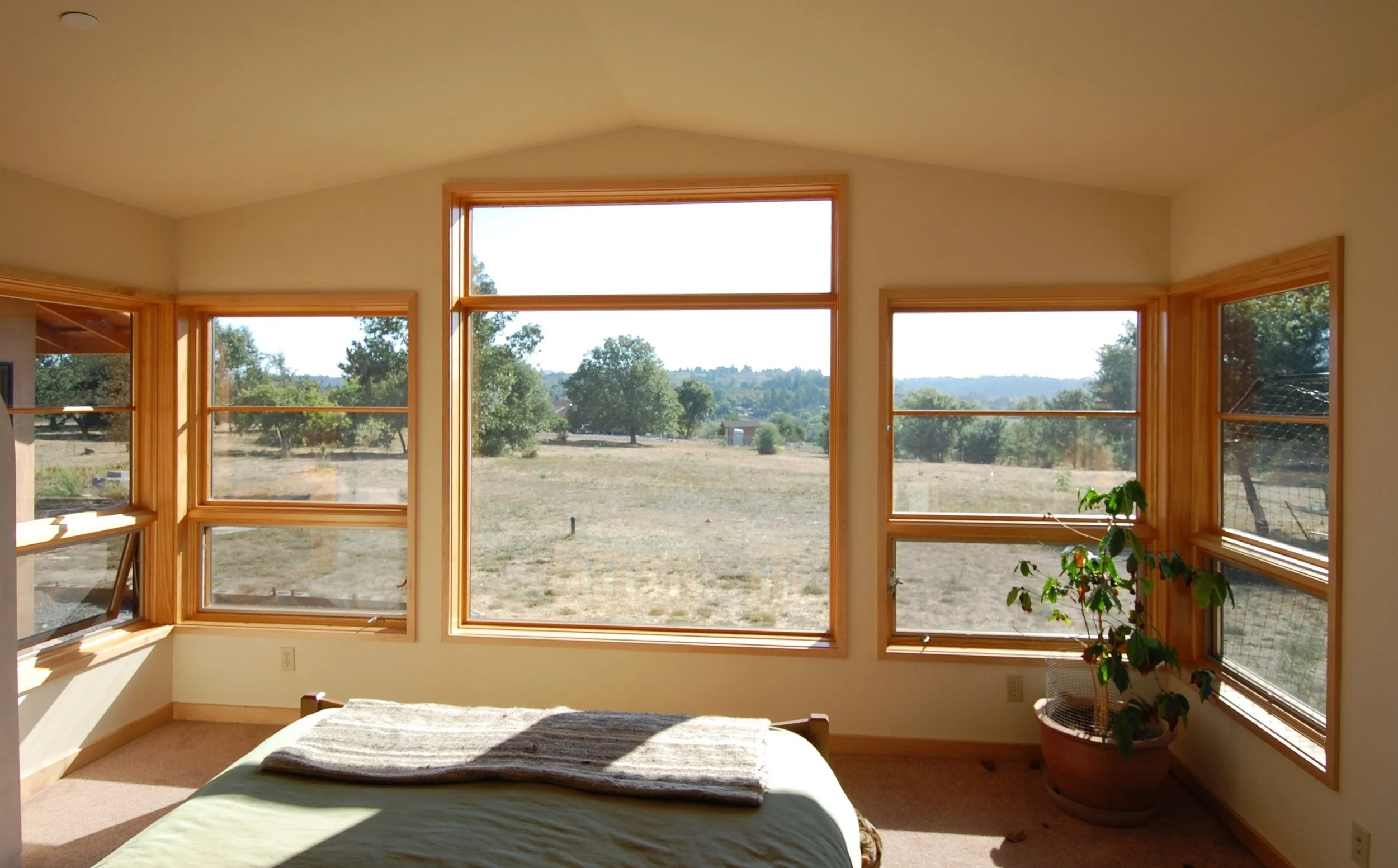 Interior of a bedroom featuring large bay windows with wood trim, overlooking an open landscape with trees and a clear sky. Part of a bed with a beige blanket and a potted plant are visible inside.