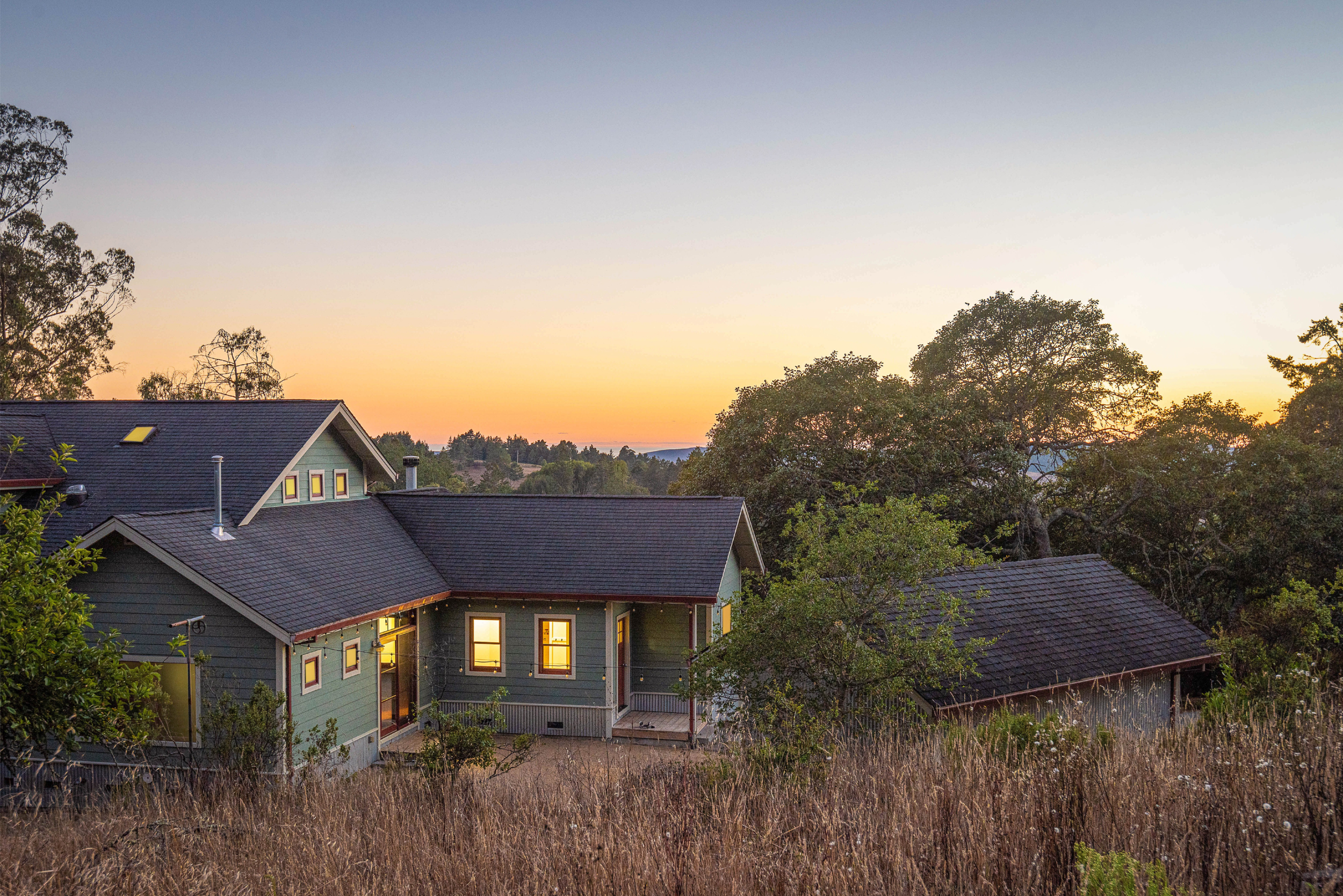 Residential houses with lit windows at sunset, trees surrounding the neighborhood, and a clear sky.
